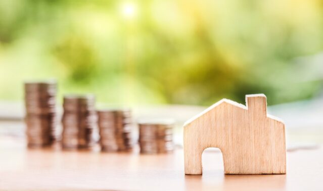 a wooden house sitting beside piles of money to represent the housing market boom